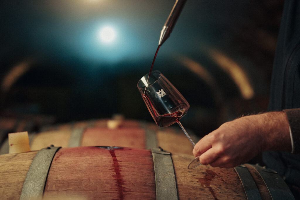 Jemand gießt Rotwein in ein Knipser-Glas. Die Gesamtszene ist stimmungsvoll in einem Weinkeller. Die Schärfe liegt auf einem Holzfass und der Einschankszene mit der Hand.
